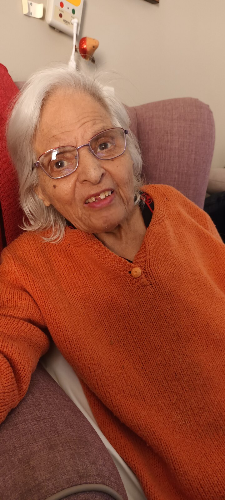 Surjit smiling cosily in her orange cardigan and glasses in her armchair