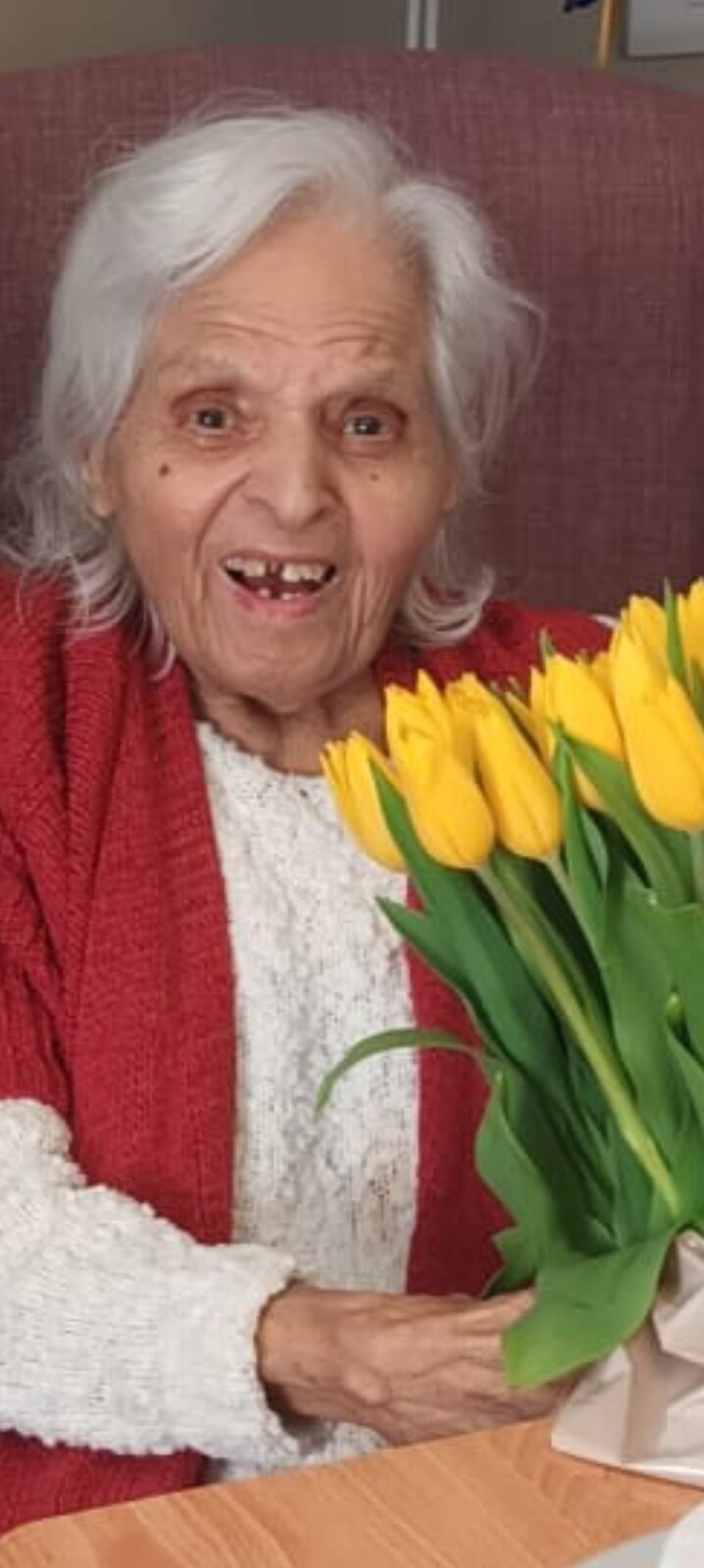 Surjit smiling warmly in a red cardigan, holding a bright bunch of yellow tulips